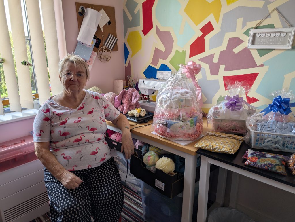 Joan, a woman with short blonde hair and glasses, sits next to her craft table which is covered in gift sets of knitted goods tied with bows.