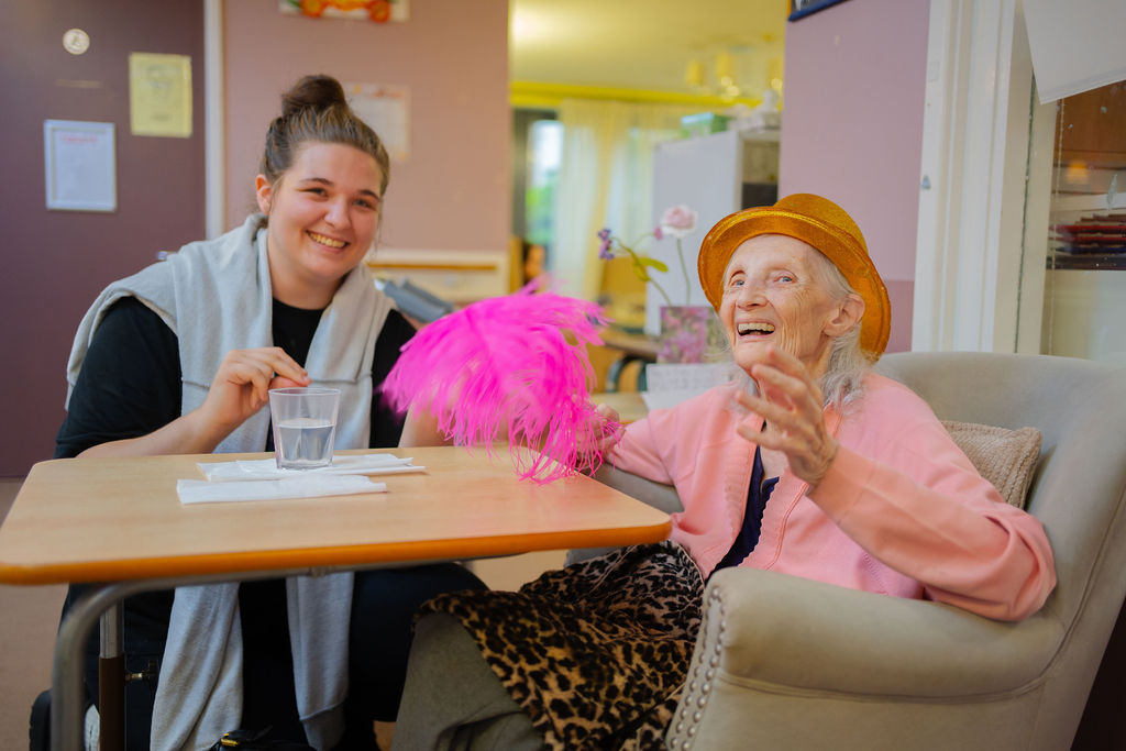 A resident wears a golds hat and holds a pom pom, and smiles with a carer.